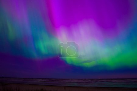 Stunning display of the Aurora Borealis over a coastal area near Riga and Jurmala, Latvia. The sky is illuminated with purple, green, and blue hues, reflecting on the calm sea.