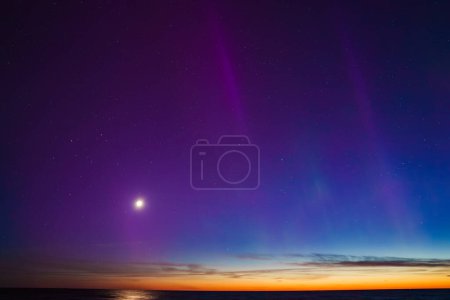 Vibrant aurora borealis illuminates the night sky near Riga and Jurmala, Latvia. The calm sea reflects hues of purple and blue, with a bright moon and setting sun.