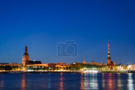 A tranquil night scene of Riga, Latvia, featuring the Daugava River reflecting city lights. Prominent landmarks include Riga Cathedral and St. Peters Church.