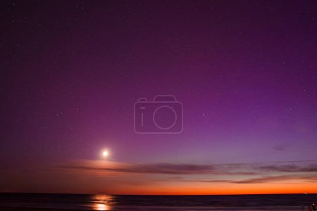 Impresionante vista de la aurora boreal con vibrantes tonos púrpura y naranja, una luna brillante que refleja la calma del mar Báltico cerca de Riga y Jurmala en Letonia.