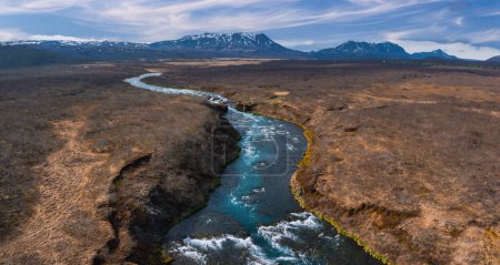 Atemberaubende Luftaufnahme eines lebendigen blauen Flusses, der sich durch eine karge Landschaft in Island schlängelt, mit schneebedeckten Bergen in der Ferne unter einem klaren blauen Himmel.