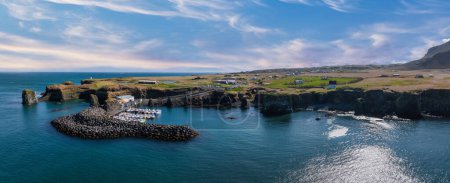 A stunning aerial view of a small harbor in Iceland, surrounded by rugged cliffs and green vegetation. The deep blue water contrasts with the bright blue sky and white clouds.