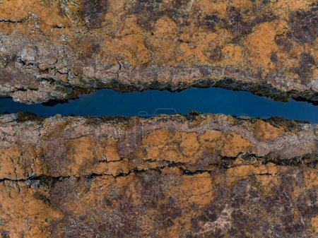 Una impresionante vista aérea de una profunda grieta en la tierra llena de un arroyo azul, rodeada por una mezcla de marrón, naranja y verde en la zona geotérmica de Islandia.