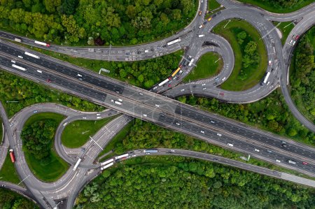Aerial view directly above a busy M62 UK motorway intersection with roundabout and overbridge cutting through green woodland in an environmental disaster image