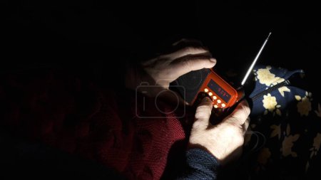 Ukraine: Grandmother holds radio in hands sitting in darkness blackout during russian terroristic attacks. Senior woman in winter clothes in darkness on sofa with hand radio receiver listening news.