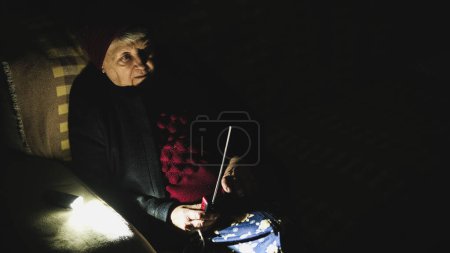 Foto de Ucrania: La abuela sostiene la radio en manos sentadas en apagón durante los ataques terroristas rusos. Mujer mayor en ropa de invierno en la oscuridad en el sofá con receptor de radio de mano escuchar noticias. - Imagen libre de derechos