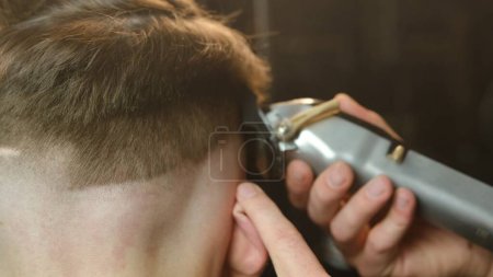 Foto de Close up men's hairstyling and haircutting with hair clipper in a barber shop or hair salon. Hairdresser service in a modern barbershop in a dark key lightning with warm light and smoke back view. - Imagen libre de derechos