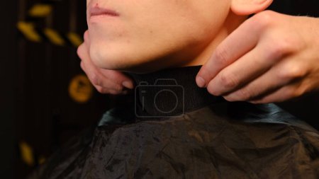 Photo for Close up Hairdresser putting on special paper collar on neck of his client before cutting hair. Hairdresser service in a modern barbershop in a dark key lightning with warm light side view. - Royalty Free Image