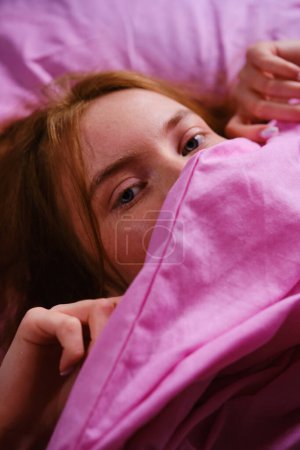 Foto de Joven pelirroja Adolescente Chica con ojos azules y pelos rojos Relajándose en la cama rosa en casa cerrando la nariz con sábana rosa jugando peekaboo. - Imagen libre de derechos