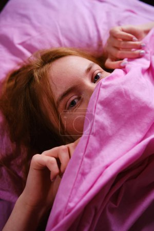 Joven pelirroja Adolescente Chica con ojos azules y pelos rojos Relajándose en la cama rosa en casa cerrando la nariz con sábana rosa jugando peekaboo.