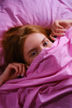 Young redhead Teenager Girl with blue eyes and red hairs Relaxing On pink Bed At Home closing her nose with pink sheet playing peekaboo.