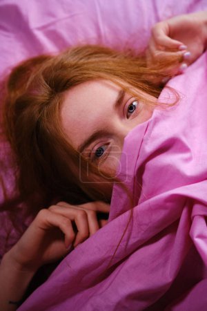 Jeune adolescente rousse aux yeux bleus et aux cheveux rouges Se détendre sur un lit rose à la maison fermant son nez avec un drap rose jouant au peekaboo.
