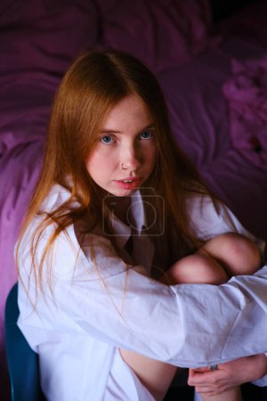 Amazing ginger woman in long white shirt sitting at the chair Fashion portrait Studio shot Beautiful red hairs girl in white at home on the pink backgtound bed hugs the legs with her hands.