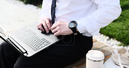 Kaukasischer bärtiger Geschäftsmann, der freiberuflich mit Laptop im Freien im Park arbeitet. Männliche Büroangestellte auf einer Bank in einem Stadtpark mit modernem urbanem Hintergrund.