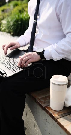 Kaukasischer bärtiger Geschäftsmann, der freiberuflich mit Laptop im Freien im Park arbeitet. Männliche Büroangestellte auf einer Bank in einem Stadtpark mit modernem urbanem Hintergrund.