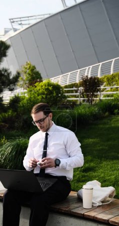 freelancer masculino que trabaja remotamente en línea con auriculares y videoconferencia portátil o reuniones de distancias Hombre en el parque de la ciudad en un banco un fondo urbano moderno, afuera, al aire libre.