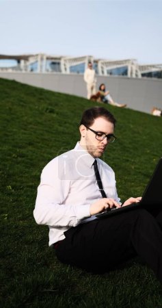 Kaukasischer bärtiger Geschäftsmann, der freiberuflich mit Laptop im Freien im Park arbeitet. Männliche Büroangestellte sitzt auf einem grünen Gras in einem Stadtpark mit modernem urbanem Hintergrund.