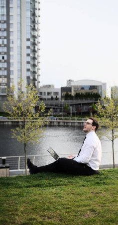 Kaukasischer bärtiger Geschäftsmann, der freiberuflich mit Laptop im Freien im Park arbeitet. Männliche Büroangestellte sitzt auf einem grünen Gras in einem Stadtpark mit modernem urbanem Hintergrund.