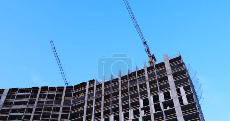 Two construction cranes tower over a multi-story building under concrete construction, windows yet to be installed, against a blue sky backdrop. Ideal for showcasing urban development.