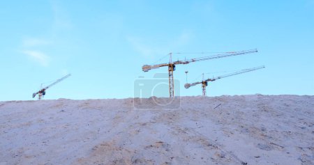 Three construction cranes tower over a mound of sand against a backdrop of blue sky. Perfect for illustrating construction progress and industrial development.