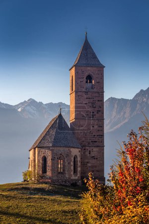 Foto de Hafling, Italia - La iglesia de montaña de Santa Catalina (Chiesa di Santa Caterina) cerca de Hafling - Avelengo en una cálida puesta de sol de otoño con los Dolomitas italianos en el Tirol del Sur al fondo - Imagen libre de derechos