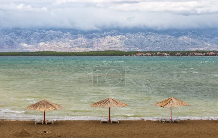 Nin, Kroatien - Drei Schilfschirme am leeren Strand der Königin bei der Stadt Nin am Ende des Sommers mit Velebit-Gebirge im Hintergrund und türkisfarbener Adria an einem bewölkten Herbsttag