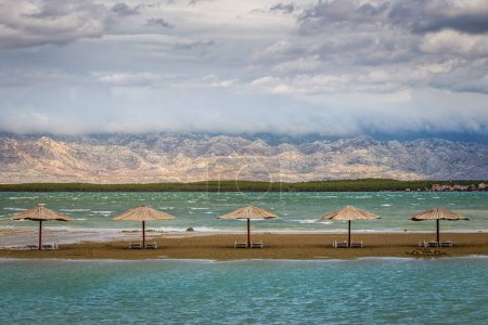 Téléchargez les photos : Nin, Croatie - Vider Queen's Beach près de la petite ville méditerranéenne de Nin à la fin de l'été avec des parasols roseaux, des montagnes Velebit à l'arrière-plan et de l'eau de mer turquoise Adriatique un jour d'automne - en image libre de droit