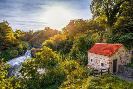 Photo for Krka, Croatia - Beautiful summer sunrise scenery of small stone house at Krka Waterfalls in Krka National Park, Dalmatia with warm morning colored foliage, rising sun and blue sky - Royalty Free Image