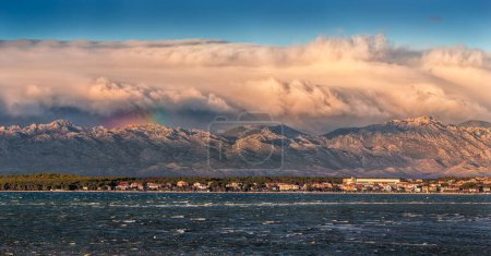 Photo for Vrsi, Croatia - Panoramic view of Vrsi municipality in Zadar county with Velebit mountains and Paklenica national park covered with warm golden colored clouds and rainbow in background at sunset - Royalty Free Image