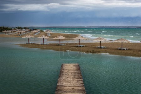 Téléchargez les photos : Nin, Croatie - Vider Queen's Beach près de la petite ville méditerranéenne de Nin à la fin de l'été avec des parasols roseaux, une petite jetée et de l'eau de mer turquoise de l'Adriatique par une journée nuageuse d'automne - en image libre de droit