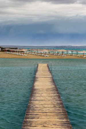 Téléchargez les photos : Nin, Croatie - Vider Queen's Beach près de la petite ville méditerranéenne de Nin à la fin de l'été avec des parasols roseaux, une petite jetée et de l'eau de mer turquoise de l'Adriatique par une journée nuageuse d'automne - en image libre de droit