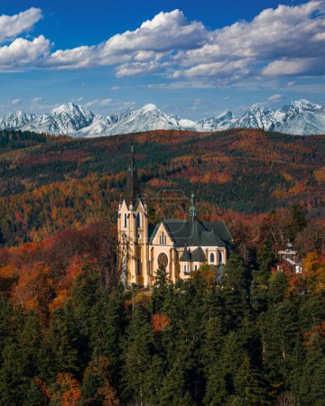 Foto de Levoca, Eslovaquia - Vista aérea de la Basílica de la Santísima Virgen María en un brillante día de otoño con la cumbre nevada de la montaña Marianska de los Altos Tatras y el cielo azul con nubes al fondo - Imagen libre de derechos
