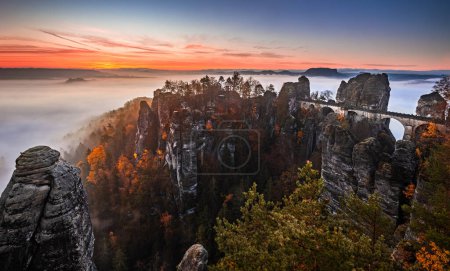 Photo for Saxon, Germany - Panoramic view of the Bastei bridge on a sunny autumn sunrise with colorful foliage and heavy fog under the valley. Bastei is famous for the beautiful rock formation in Saxon Switzerland National Park near Dresden - Royalty Free Image
