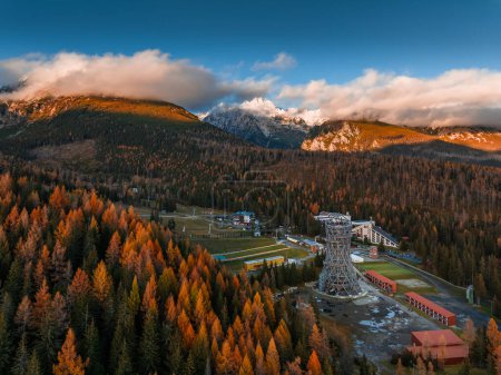 Photo for Strbske Pleso, Slovakia - Aerial view of the Strbske Lake area with autumn foliage, sightseeing tower and the High Tatras mountains at background on an autumn afternoon at sunset with warm sunlight, blue sky and clouds - Royalty Free Image