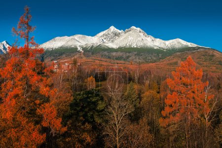 Tatranske Matliare, Slowakei - Luftaufnahme der schneebedeckten Berge der Lomnitzspitze in der Hohen Tatra mit schönen rot und orange gefärbten Herbstbäumen und Laub und klarem blauen Himmel in der Vysoke Tatry