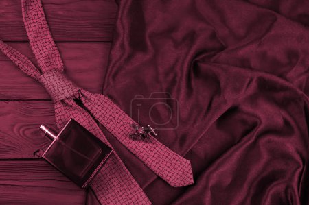 A bottle of mens cologne and cufflinks with blue tie lie on a black luxury fabric background on a wooden table. Mens classic accessories. Shallow DOF Image toned in Viva Magenta, color of the year