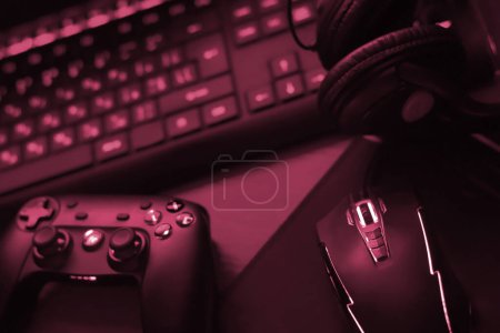 Photo for Modern gamepad and gaming mouse lies with keyboard and headphones on table in dark playroom scene close up. Video game challenges and competitions Image toned in Viva Magenta, color of the 2023 year - Royalty Free Image