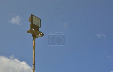 Foto de Una cámara CCTV y un proyector cuadrado están montados en un poste de metal contra el cielo azul. Sistema de videovigilancia organizado - Imagen libre de derechos