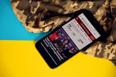 Foto de KYIV, UCRANIA - 4 MAYO 2023: UP Ukrainska Pravda portal de noticias ucraniano en la pantalla del teléfono inteligente con bandera ucraniana y tela de camuflaje de cerca - Imagen libre de derechos