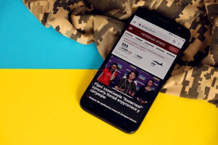 Foto de KYIV, UCRANIA - 4 MAYO 2023: UP Ukrainska Pravda portal de noticias ucraniano en la pantalla del teléfono inteligente con bandera ucraniana y tela de camuflaje de cerca - Imagen libre de derechos