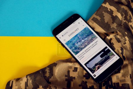 Photo for KYIV, UKRAINE - 4 MAY, 2023: Zaxid Net ukrainian news portal on smartphone screen with ukrainian flag and camouflage fabric close up - Royalty Free Image
