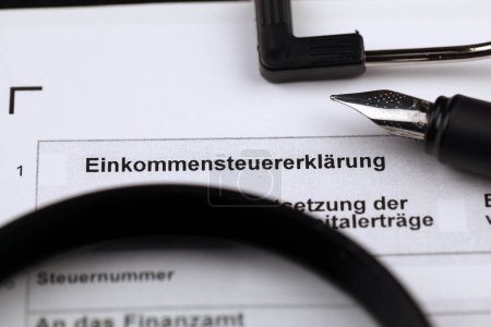 Deutsches Formular zur jährlichen Einkommensteuererklärung blanko auf DIN-A4-Tablet liegt mit Stift und Lupe in Großaufnahme auf dem Bürotisch