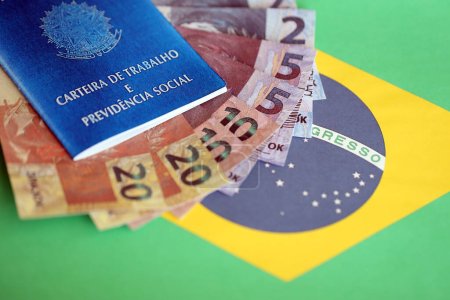 Photo for Brazilian work card and social security blue book and reais money bills on flag of Federative Republic of Brazil close up - Royalty Free Image