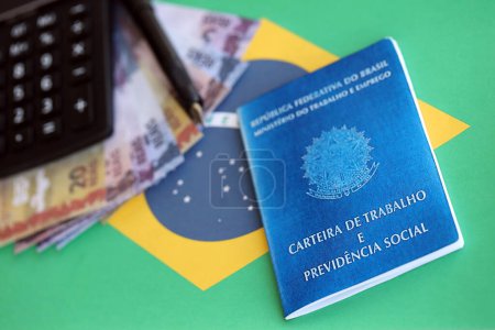 Brazilian work card and social security blue book and reais money bills with calculator and pen on flag of Federative Republic of Brazil close up
