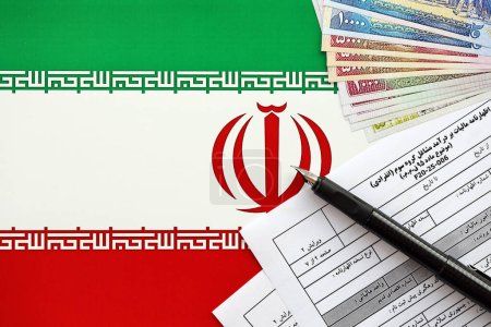 Iranisches Formular für die jährliche Einkommensteuererklärung F20-25-006 fertig zum Ausfüllen auf dem Tisch mit Stift und iranischem Geld auf der Fahne in Großaufnahme
