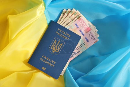 One Ukrainian biometrical passport and UAH hryvnia bills on folded waving flag of Ukraine country close up