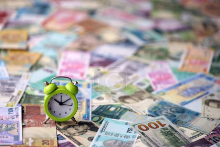 Small alarm clock on many banknotes of different currency. Background of time and money close up