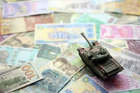Small green tank on many banknotes of different currency. Background of war funding or spend money to defense close up