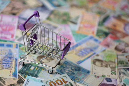 Supermarket trolley on many banknotes of different currency. Background of shopping worldwide close up