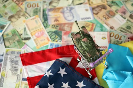 Tanque de juguete en bandera estadounidense y bandera ucraniana en muchos billetes de diferentes monedas. Antecedentes de financiación de la guerra y apoyo militar entre Estados Unidos y Ucrania